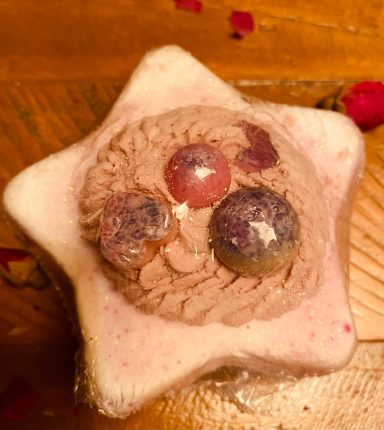 A star shaped bath bomb with pink base, lilac bubble icing and 2 round cocoa butter melts that are pink and purple in colour