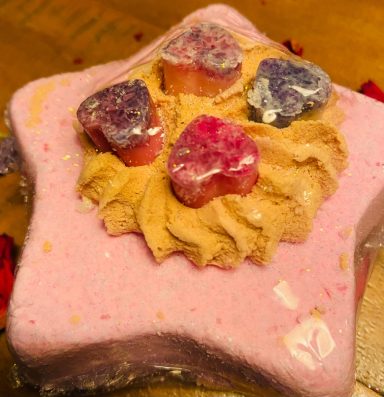 A pink star shaped bath bomb with gold bubble icing piped on top and 4 small cocoa butter heart melts on top these are pink and purple